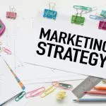 Tulisan "marketing strategy" di atas kertas dengan kalkulator, buku, dan alat tulis